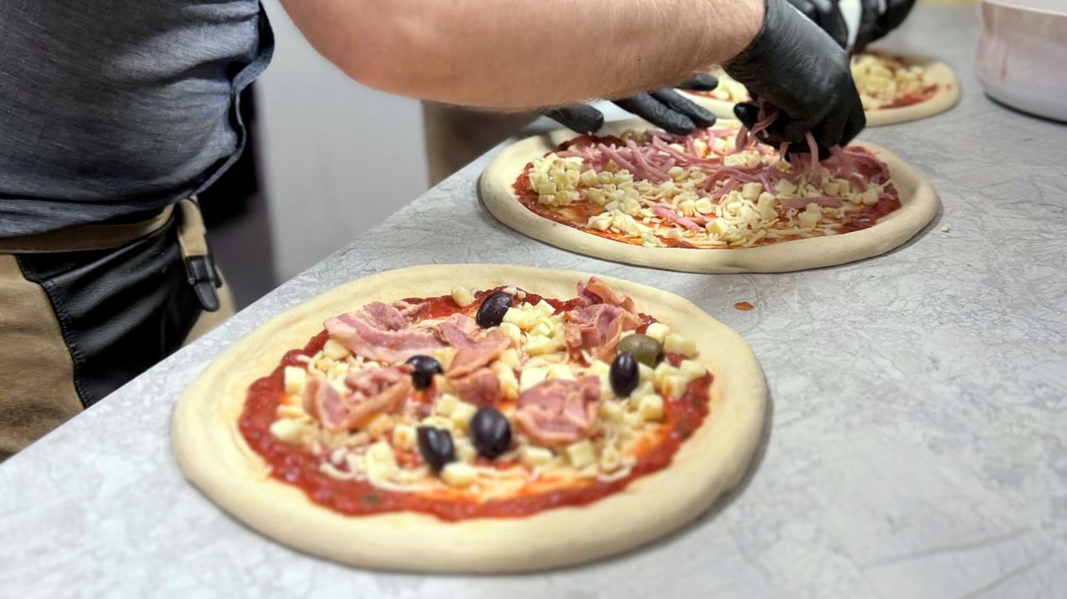 Två kockar lägger skinka och ost på pizzor i köket